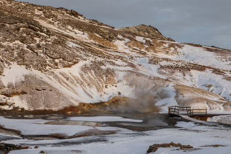 Snow covered geothermal hills rise above shallow pools at Seltun Krysuvik in Iceland. Warm light highlights the mixed volcanic and snowy terrain.の写真素材
