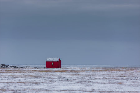 A small red cabin stands alone in a vast snowy field near Krysuvik in Iceland. Low clouds create a muted winter atmosphere over the open landscape.の写真素材