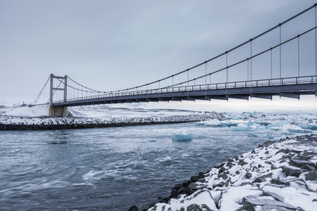 A suspension bridge spans the outflow channel of Jokulsarlon Glacier Lagoon in Iceland, with floating icebergs and snow-covered mountains in the background.の写真素材