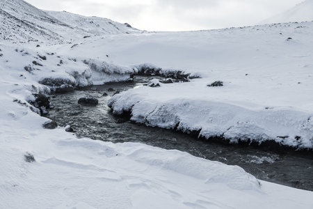 Geothermal hot spring stream flowing through a snow covered Reykjadalur Valley in Iceland, surrounded by winter hills and frozen volcanic landscape.の写真素材