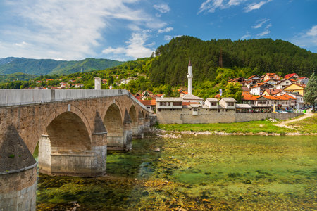 Konjic, Bosnia and Herzegovina â August 16, 2022: Old stone bridge over the Neretva River with historic riverside buildings, mosque minaret and hillside houses in the town of Konjic.のeditorial素材