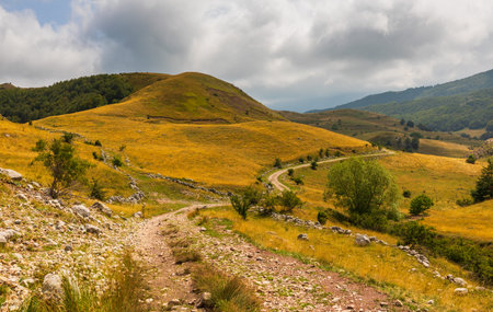 Lukomir, Bosnia and Hercegovina - August 23, 2022: Winding road traversing dry grass hills under a cloudy sky.のeditorial素材