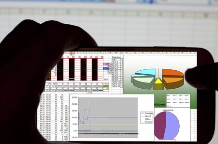 Silhouetted male hands holding a smartphone and using a spreadsheet application in the officeの写真素材
