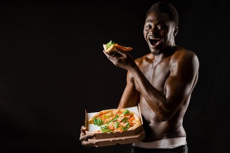 Black handsome naked man eats a pizza. Safety food delivery for people who stays at home. Sexy african man shows 2 pieces of pizza in his hands. Tasty food from pizzeria restaurantの写真素材