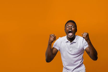 Happy handsome black man wins online bet on orange background. Shouting loud "Yes" and making crazy emotions. African won a prize. Online work at home quarantine concept. Empty left side for advertの写真素材
