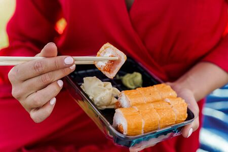 Close-up sushi set. Attractive girl in red dress holds sushi with chopsticks in hands. Courier delivered sushi set for pretty woman. Black box with sushi, wasabi, ginger.の写真素材