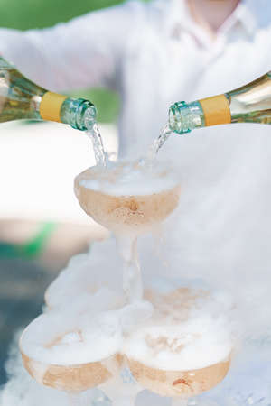 Waiter pours champagne from 2 bottles into crystal glasses with dry ice and white smoke close up. Sparkling wine pouring in wine glass at summer day. Catering for business people. Vertical photo.の写真素材