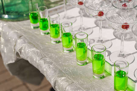 Shot glasses with green syrup near pyramid of glasses with champagne and red cherry inside on...