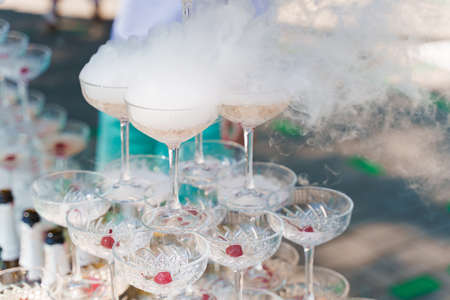 The waiter pours sparkling wine into crystal glasses with dry ice and white smoke close up. Champagne pouring in wine glass at summer day. Catering for wedding ceremony.の写真素材