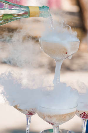 The waiter pours champagne into crystal glasses with dry ice and white smoke close up. Sparkling wine pouring in wine glass at summer day. Catering for business people. Vertical photo.の写真素材