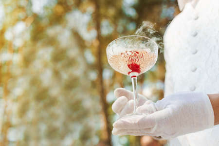 Waiter in white gloves gives wine glass with shampagne, red cherry, and white smoke of dry ice and gives to customer. Catering for wedding ceremony and business meeting. Empy left side for text.の写真素材
