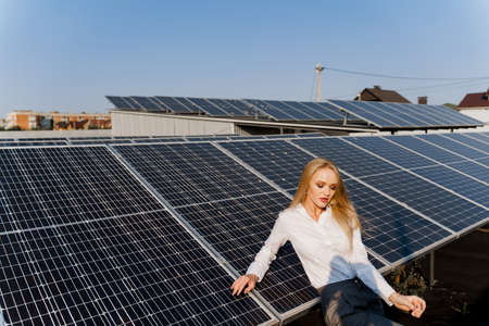 Woman leans on solar panels. Blonde dressed white formal shirt on the power plant. Free electricity for home. Green energy. Solar cells power plant business.の写真素材