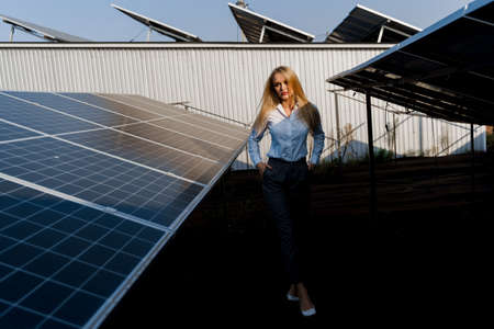 Girl walks between 2 Solar panels row on the ground at sunset. Woman investor wears formal white...