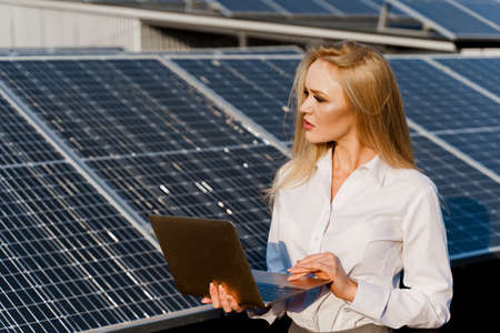 Investor woman stand with laptop near blue solar panels row on the ground Girl weared formal white...
