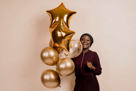 Christmas celebration. African american holiday. Attractive girl with golden helium balloons isolated peach background. Young black woman weared traditional dress and scarf.の写真素材