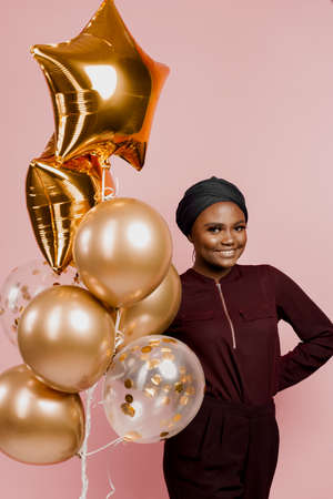 Christmas celebration. African american holiday. Attractive girl with golden helium balloons isolated peach background. Young black woman weared traditional dress and scarf.の写真素材