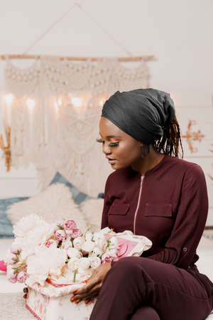 Muslim girl with flowers seats and looks at bouquet. African young woman with beautiful flowers from...