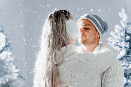 Happy young couple hugs and smiles and snow falls near christmas trees at the eve of new year celebration in winter day. Smiley man and woman weared white pullovers love each otherの写真素材