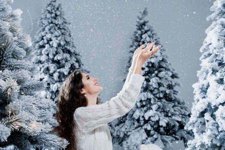 Happy girl smiles and touches falling snow at eve new year. Young woman near big snowy spruces in winter day before christmas celebrationの写真素材