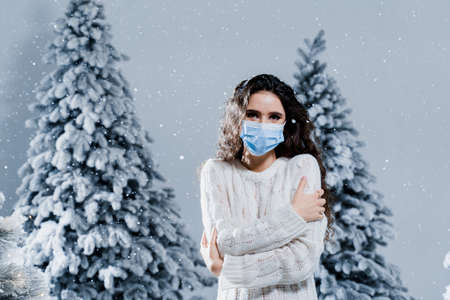 New year celebration at covid-19 coronavirus quarantine period. Happy girl in medical mask with falling snow stays at home. Social distance. Winter holidays in snowy dayの写真素材