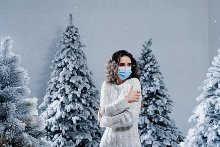 New year celebration at covid-19 coronavirus quarantine period. Happy girl in medical mask with falling snow stays at home. Social distance. Winter holidays in snowy dayの写真素材