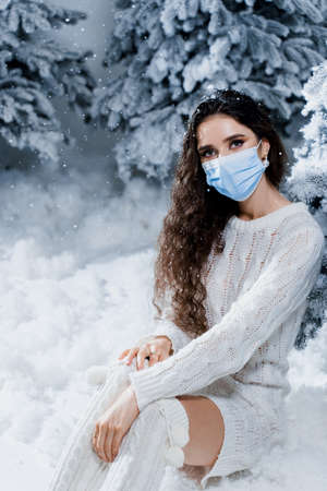 New year celebration at covid-19 coronavirus quarantine period. Happy girl in medical mask with falling snow stays at home. Social distance. Winter holidays in snowy dayの写真素材