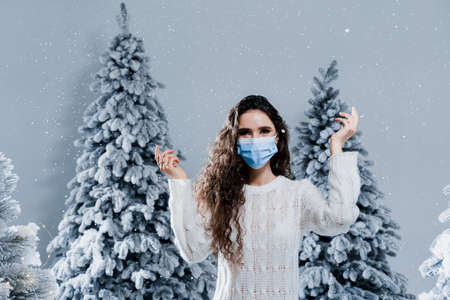 New year celebration at covid-19 coronavirus quarantine period. Happy girl in medical mask with falling snow stays at home. Social distance. Winter holidays in snowy dayの写真素材