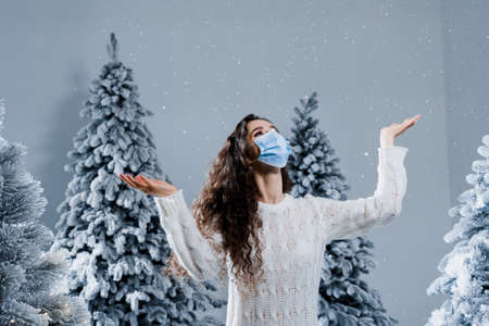 New year celebration at covid-19 coronavirus quarantine period. Happy girl in medical mask with falling snow stays at home. Social distance. Winter holidays in snowy dayの写真素材