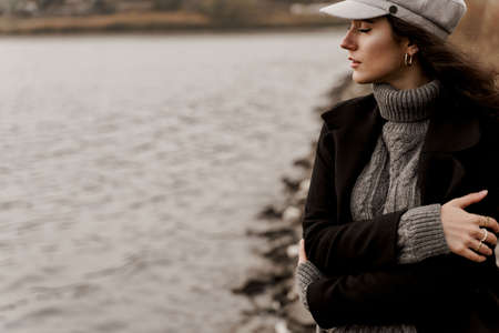A beautiful girl in a black coat and a gray woolen cap near the lake. Traveling in your country