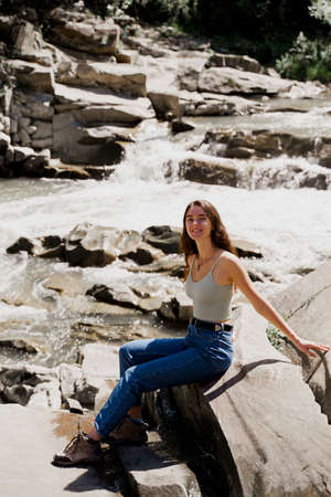 Traveler girl is sitting on the rock near waterfall and looking toward. Traveling in Karpathian mountains. Cascade waterfall. Beautiful landscapeの写真素材