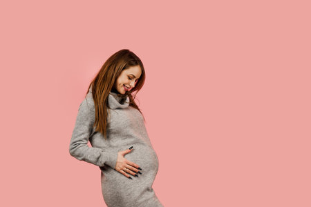 Pregnant woman. Expectation of the child. Maternity leave. Pregnancy of a happy beautiful girl in a gray sweater on a pink backgroundの写真素材