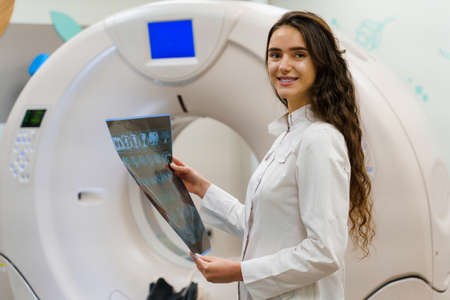 Medical student looks in camera and smile. Medical stands with x-ray picture of patient brain in her hands. woman stays in front of kt komputer tomograhphy. Girl in medical clinic gotes new expirienceの写真素材