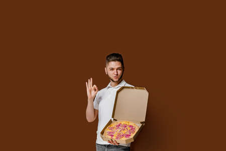 Online pizza safety delivery servise from restaurant. Young caucasian courier man shows ok and smile. Pizza with cheese boards advertise. Isolated brown background with empty space for advertiseの写真素材