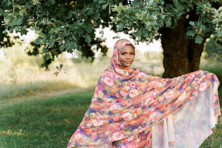 Muslim black woman African ethnicity weared traditional colorful hijab smiles under green tree. Arabic islam religion clothes. Pretty black girl smiles. Business for black woman.の写真素材
