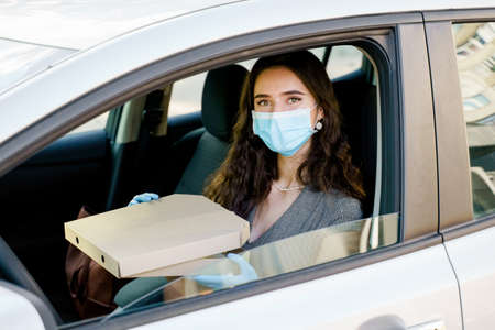 Business woman with pizza box in car. Delivery food service drive by car. Safe food delivered according social distance. Tasty pizza with salami, cheese, tomatoes in cardoard box.の写真素材