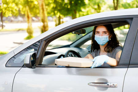 Safe food delivery from pizzeria to car during quarantine. Attractive business woman in medical mask...