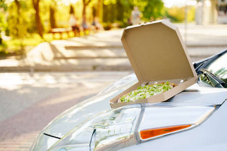 Pizza closeup on the eco car. Safe delivery of pizza with green salad, tomatoes, cheese. Advertise in social network for restaurant delivery. Ecological delivering by electric carの写真素材