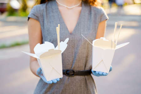 Closeup tasty food wok in box delivery. Girl holds udon noodles with tempuru, shrimps, soy sauce in hands in medical gloves. Chinese spicy food in disposable eco boxes. 2 Wok box udon advertise 1 + 1.の写真素材