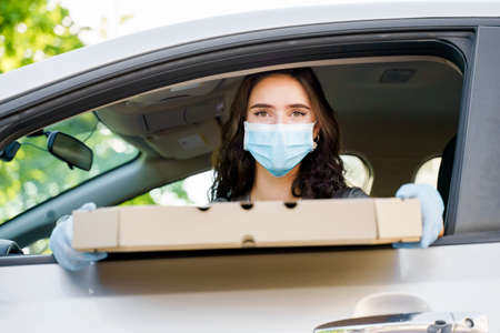 Lovely caucasian girl with green eyes looks into camera and smiles. Young girl holds pizza in car and works as courier in delivery company at quarantine covid 19. Delivering pizza by carの写真素材