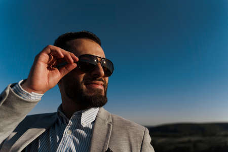 Handsome bearded arabian man in sunglasses on the blue sky background. Confident muslim business man. Arab man weared in gray coatの写真素材