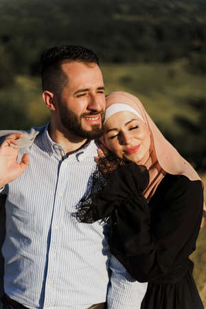 Muslim love story close-up. Mixed couple smiles and hugs on the green hills. Woman weared in hijab looks to her man. Advert for on-line dating agencyの写真素材