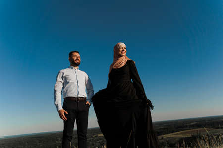 Muslim love story on the blue sky background. cheerful mixed couple smiles and hugs. Woman weared in hijab looks to her man. Advert for on-line dating agencyの写真素材