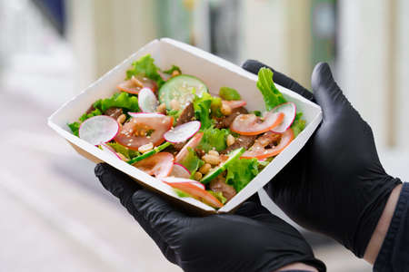 Green salad with veal and vagatables. Safety delivery with mask and black medical gloves at quarantine. Salad in eco thermo box. On line order from the restaurant delivered to people who stay at home.の写真素材
