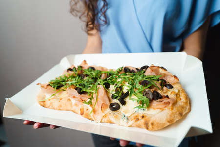 Pinsa romana in white delivery box. Courier holding crocchiarella gourmet italian cuisine on white background. Food delivery from pizzeria. Pinsa with meat, arugula, olives, cheese.の写真素材