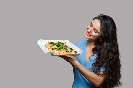 Girl courier with pinsa romana gourmet italian cuisine on gray background. Holding scrocchiarella traditional dish. Food delivery from pizzeria. Pinsa with meat, arugula, olives, cheese.の写真素材
