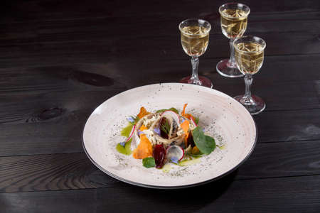 Salad of herring, mayonnaise, onion, garnished with pumpkin chips, blue onions, flowers on a white plate on a dark wooden background with stacks of alcohol.の写真素材