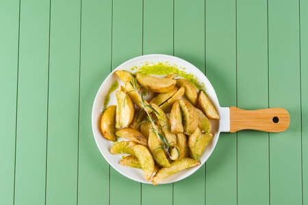 Baked potatoes with green sauce and rosemary on a white plate in the form of a frying pan. Fried potatoes with pesto sauce on a green table.の写真素材
