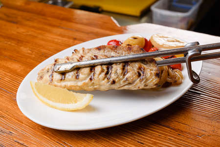 Chicken fillet and grilled vegetables with lemon on a white plate on a wooden table. Serving the...