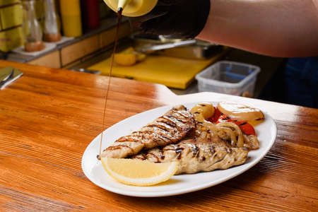 Chicken fillet and grilled vegetables with lemon on a white plate on a wooden table. Pouring sauce...
