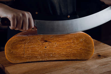 Slicing aged cheese parmesan with crystals using a cheesy dutch knife. Hard cheese with knife on dark background. Snack tasty piece of food for appetizerの写真素材